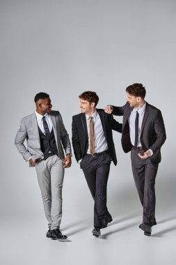 good looking cheerful multiethnic male models in smart suits smiling sincerely on gray backdrop clipart