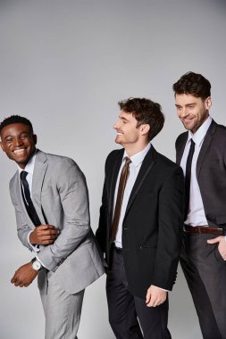 good looking cheerful multicultural male models in smart suits smiling sincerely on gray backdrop clipart