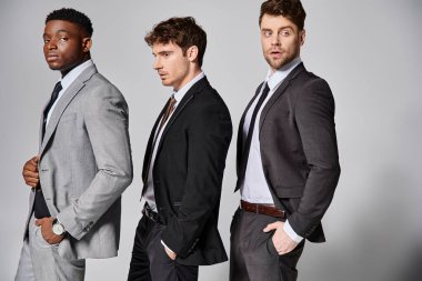 attractive stylish multicultural male models in business casual attires posing on gray backdrop clipart