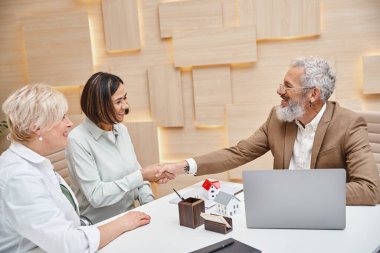 Emlakçı, emlak bürosundaki lezbiyen ortağının yanında orta yaşlı, çok ırklı bir kadınla tokalaşıyor.