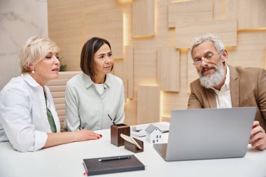 realtor showing laptop with house projects to middle aged lgbt couple in real estate office clipart