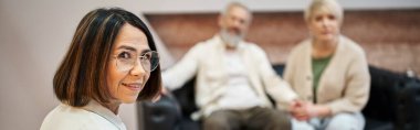 Pankart, gözlüklü orta yaşlı psikolog arka planda evli çiftin yanında kameraya bakıyor.