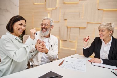 happy married couple holding key from new house near smiling realtor in real estate office clipart