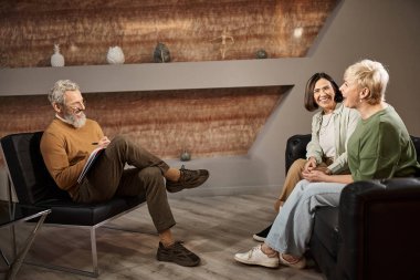 happy middle aged psychologist in glasses listening to smiling lesbian couple during therapy session clipart