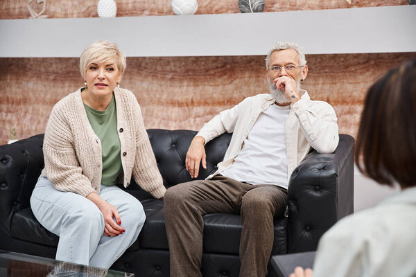 middle aged couple sitting on leather couch and looking at psychologist during consultation