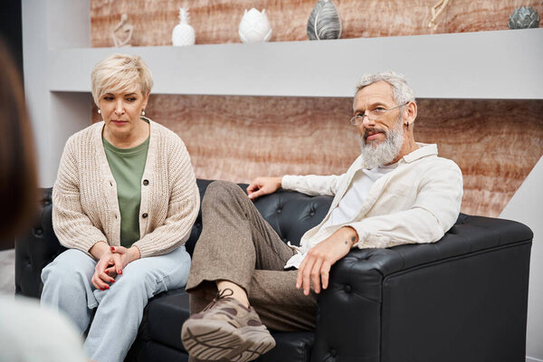 middle aged couple sitting on leather couch and looking at family counselor during consultation