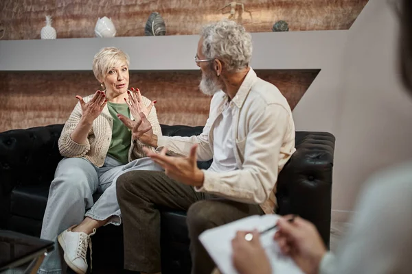 Stock image emotional middle aged couple arguing while sitting near psychologist during therapy session