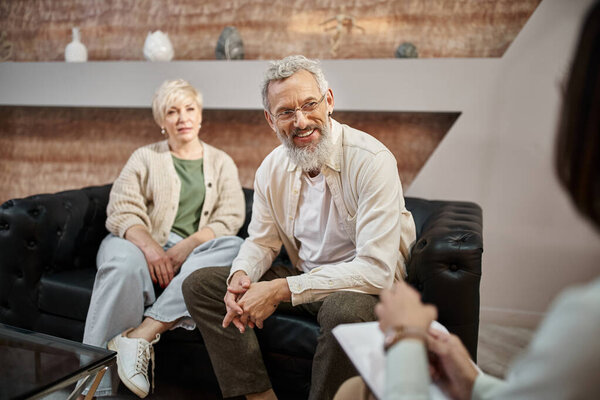 cheerful bearded man sitting on couch and looking at family consultant near wife during session