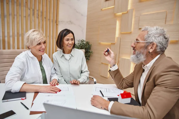 stock image bearded realtor holding key to new house near cheerful middle aged lesbian couple, real estate