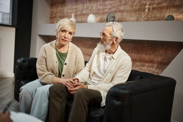 married middle aged couple holding hands and sitting on couch during family therapy session