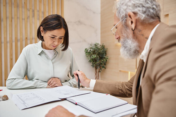 bearded realtor with tattoo pointing with pen at blueprint near woman buying real estate property
