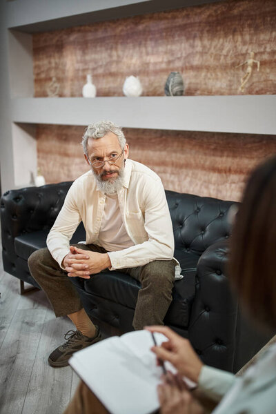 bearded middled aged man with tattoo sitting on leather couch and listening to female psychologist