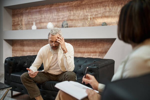 bearded middled aged man with tattoo sitting on leather couch and talking to female psychologist