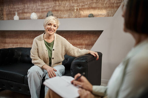 cheerful blonde middled aged woman sitting on leather couch and looking at psychologist on session