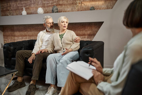 bearded man hugging worried middle aged wife looking at psychologist during therapy session