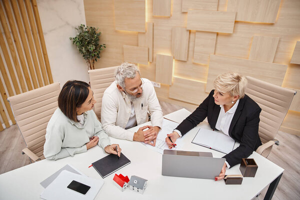 middle aged realtor showing project of new house on laptop to married couple in real estate office