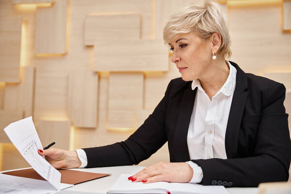 блондинка среднего возраста риэлтор с короткими волосами работает в офисе недвижимости, глядя на чертеж