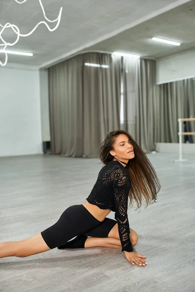 flexible african american dancer in black clothes rehearsing on floor in modern dance studio