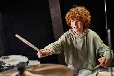 cheerful adorable talented teenage boy in casual outfit playing his drums actively in studio clipart
