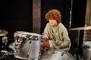 concentrated cute red haired teenage boy in casual comfy clothes playing his drums in studio clipart