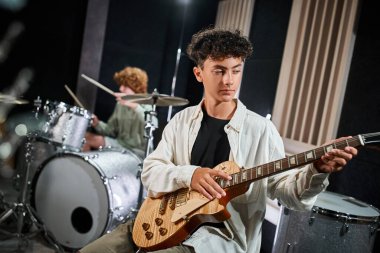 concentrated adorable teenage boy in casual attire playing guitar next to his blurred drummer clipart