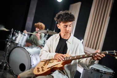 focused talented teens in casual attires playing guitar and drums in studio, musical group clipart