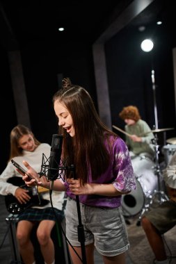 cheerful teenage girl in vivid attire singing and looking at her mobile phone next to her friends clipart