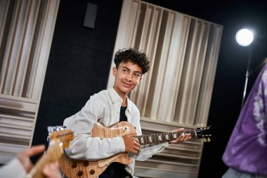 Güzel görünümlü, sıradan giyinmiş gitar çalan ve yetenekli arkadaşlarına bakan genç bir çocuk.