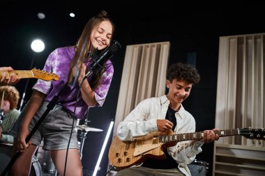 adorable joyful teenage girl singing happily while her friend playing instruments in studio clipart