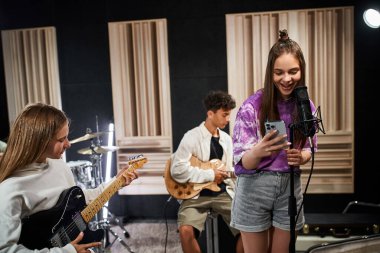 Neşeli genç kız telefona bakıyor ve arkadaşlarının yanında şarkı söylüyor gitar çalıyor, müzik grubu