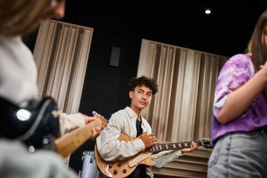 Gitar tutan ve stüdyodaki bulanık müzik grubu üyelerine bakan sevimli bir çocuk.