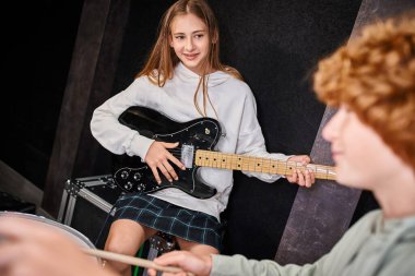 focus on jolly pretty teenage girl holding her guitar and looking at her blurred cute drummer clipart