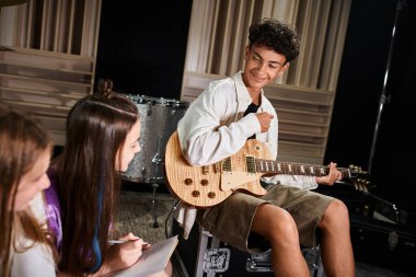 Gitar tutan ve bulanık müzik grubu üyelerine bakan neşeli, genç bir çocuğa odaklan.
