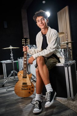 Gitar tutan ve stüdyoda kameraya gülümseyen sıradan giyinmiş neşeli, yakışıklı genç bir çocuk.
