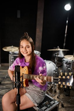 Canlı giyinmiş, gitar çalan ve kameraya bakan neşeli genç gitarist.