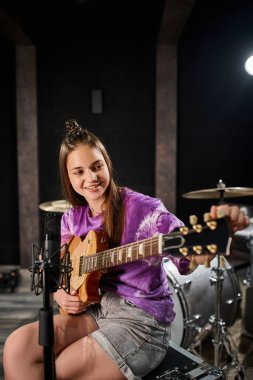 Canlı giyinmiş neşeli genç gitarist stüdyoda mikrofonun önünde gitar çalıyor.