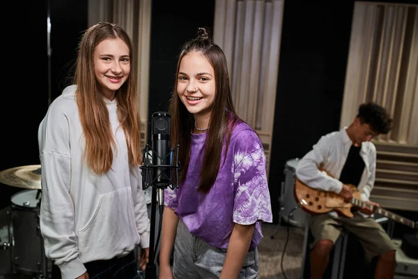 stock image joyful pretty teenage girls looking at camera near microphone with their friend playing guitar