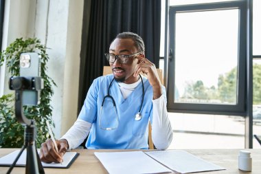 Neşeli, yakışıklı, Afro-Amerikan doktor telefonla konuşurken not alıyor.