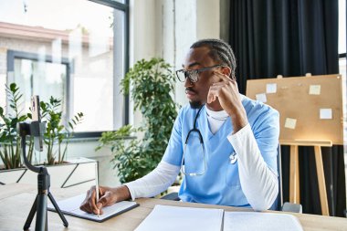 Düşünceli yakışıklı Afrikalı Amerikalı doktor telefonla konuşurken not alıyor, teletıp...