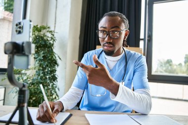 Odaklanmış yakışıklı Afro-Amerikan doktor telefonla konuşurken not alıyor, teletıp