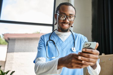 handsome cheerful african american doctor with stethoscope looking at his mobile phone, telemedicine clipart