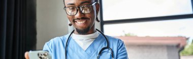 Steteskoplu neşeli Afrikalı Amerikalı doktor cep telefonuna, teletıp ve afişine bakıyor.