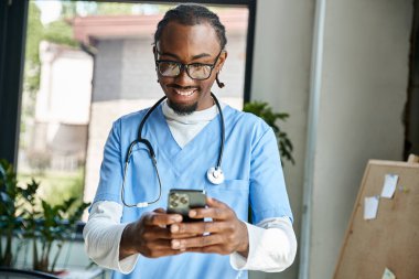Steteskoplu yakışıklı Afro-Amerikan doktor cep telefonuna bakıyor, teletıp.