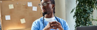 Steteskoplu düşünceli Afrikalı Amerikalı doktor ofisinde oturuyor ve başka tarafa bakıyor.