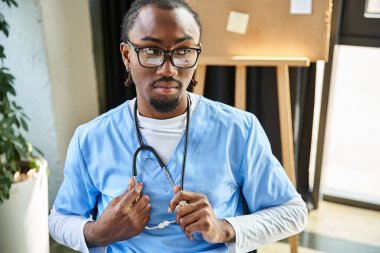 Düşünceli, yakışıklı, gözlüklü Amerikalı Amerikalı doktor steteskop tutuyor ve başka tarafa bakıyor.