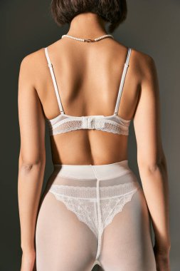 Beyaz dantelli iç çamaşırlı, külotlu çoraplı, gri arka planda poz veren genç bir kadın.
