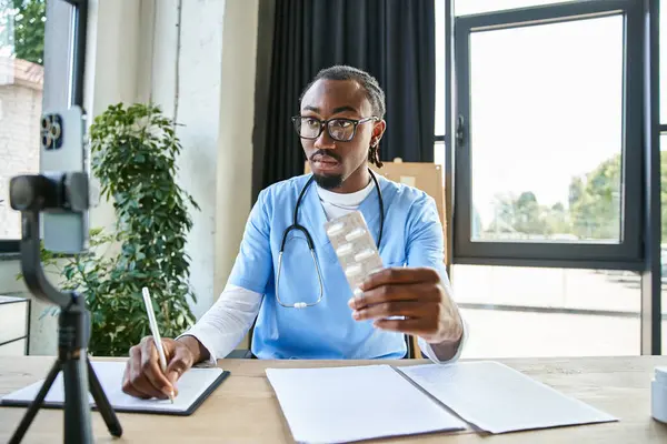 stock image concentrated african american doctor holding pills and taking notes while consulting by phone