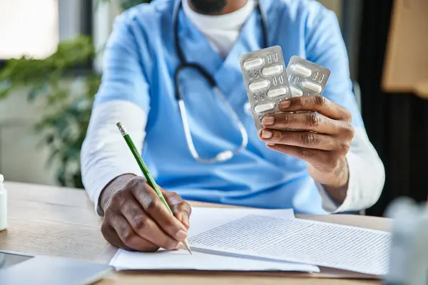 stock image cropped view of african american doctor with stethoscope holding pills and taking notes, telehealth