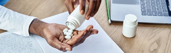 stock image cropped view of young african american doctor pouring pills into his hand next to laptop, banner