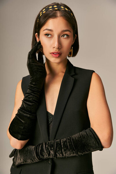 charming asian woman in elegant attire with gloves and headband posing on grey background